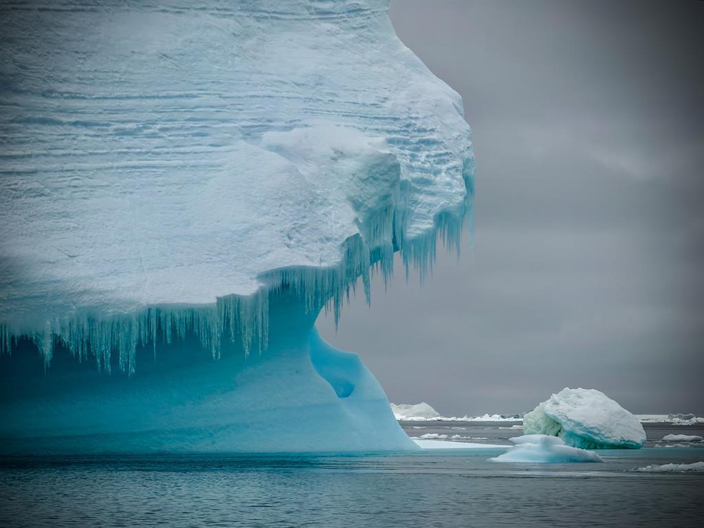 Links ragt der Eisfels aus dem Meer empor. Die Farben des Fotos sind grau, blau, schwarz und weiß.